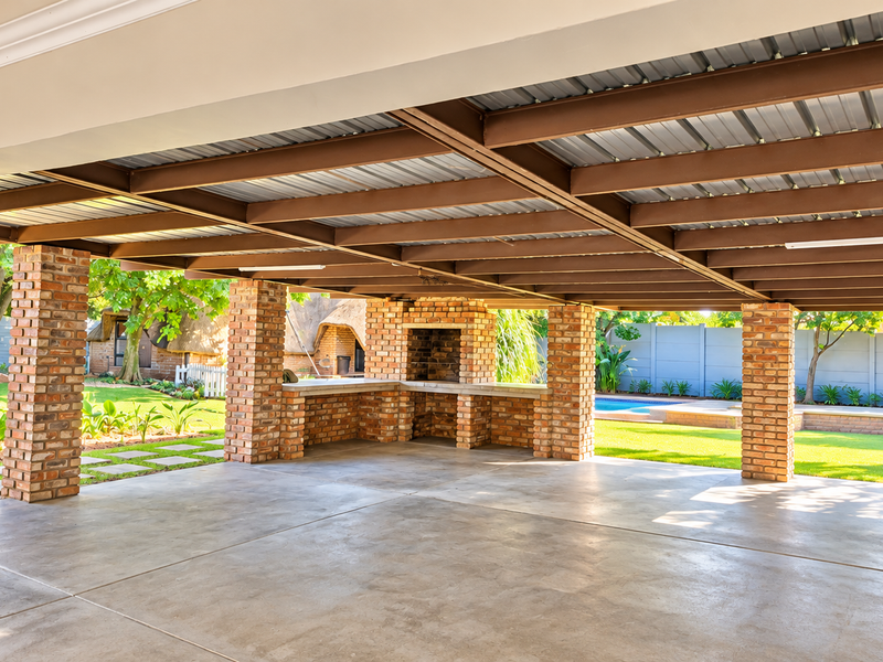 Spacious covered outdoor patio featuring exposed steel beams, brick columns, and a built-in brick braai, overlooking a landscaped garden and pool area.