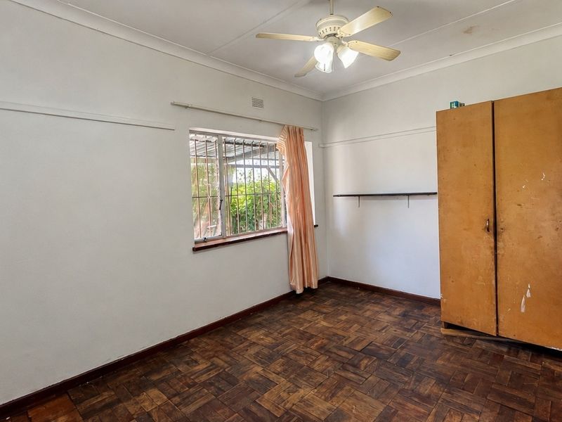 A bedroom featuring original parquet flooring, a ceiling fan, and security bars on the window, offering a blank canvas for renovation.