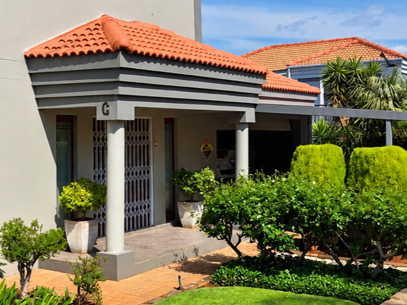 Front elevation of a townhouse featuring a covered entrance porch with concrete columns, terracotta roof tiles, and a well-maintained landscaped garden.