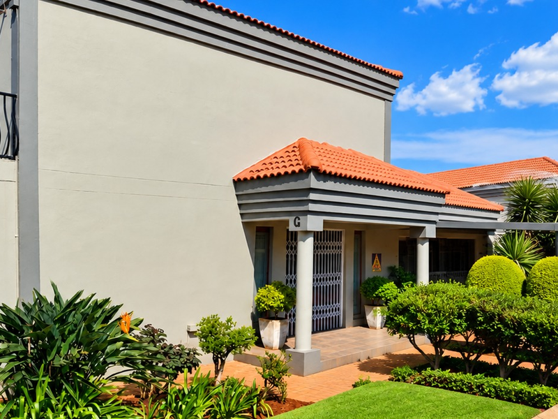 A well-maintained townhouse exterior featuring a covered entrance, terracotta roof tiles, and neatly landscaped garden beds with paved walkways.
