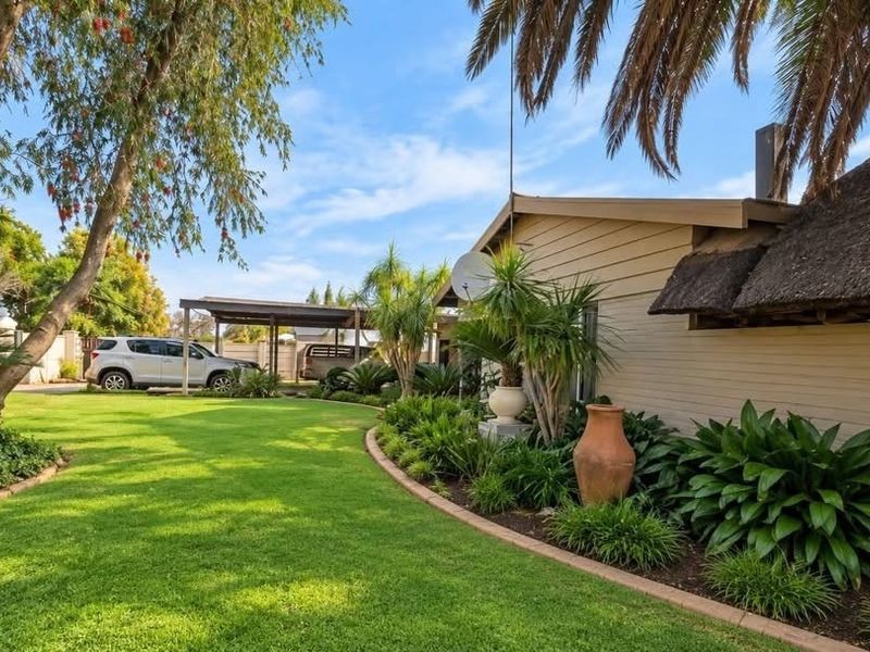 A well-maintained rear garden featuring a lush lawn, mature trees, landscaped garden beds, and a section of the house with a traditional thatched roof.