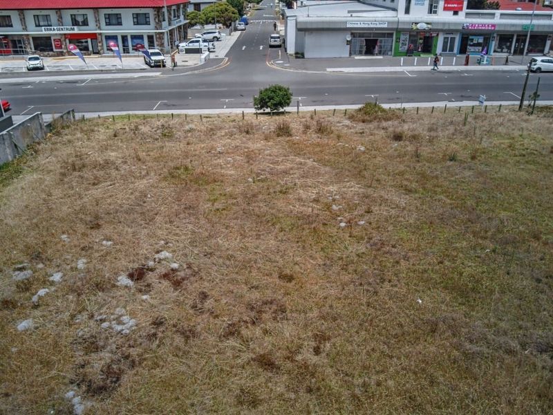 Aerial view of a vacant commercial land plot situated in a high-visibility street location, featuring boundary fencing and proximity to local retail infrastructure.