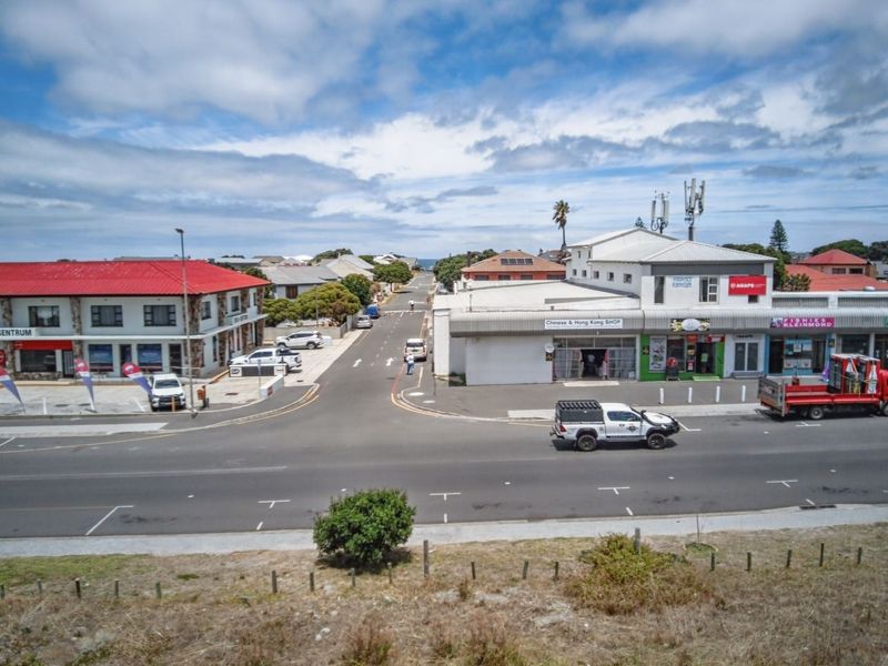 Aerial view of a commercial street intersection featuring established retail buildings, paved parking, and road infrastructure in a coastal town setting.