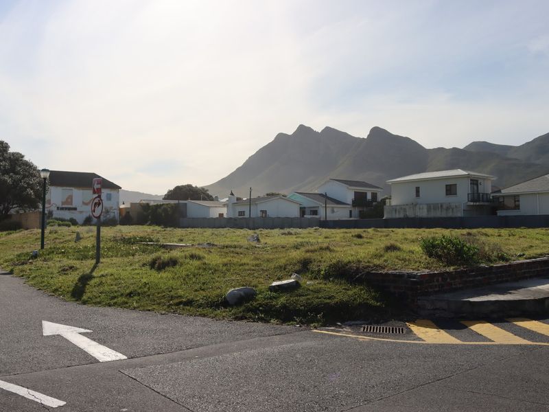 A vacant commercial land plot situated on a corner, featuring grassed terrain, a brick retaining wall, and scenic mountain views in the background.