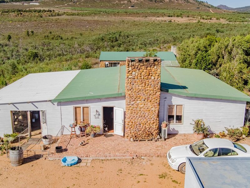 Aerial view of a rural property featuring a single-story building with a prominent stone chimney, metal roofing, and an adjacent brick patio area.