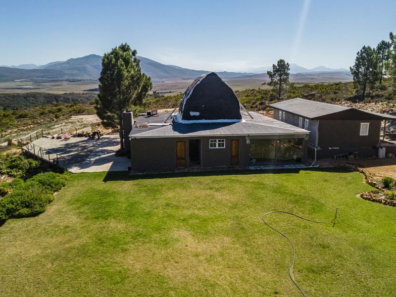Aerial view of a farm property featuring a unique dome-roof structure, expansive lawn, and a paved patio area set against a scenic mountain landscape.