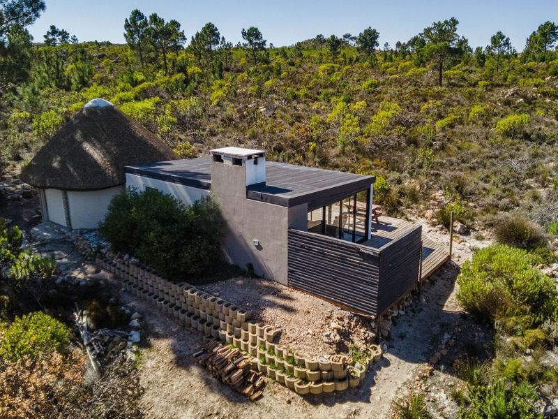 Aerial view of a modern farm dwelling featuring a thatched roof section, flat roof extension, and a timber deck set within a natural landscape.