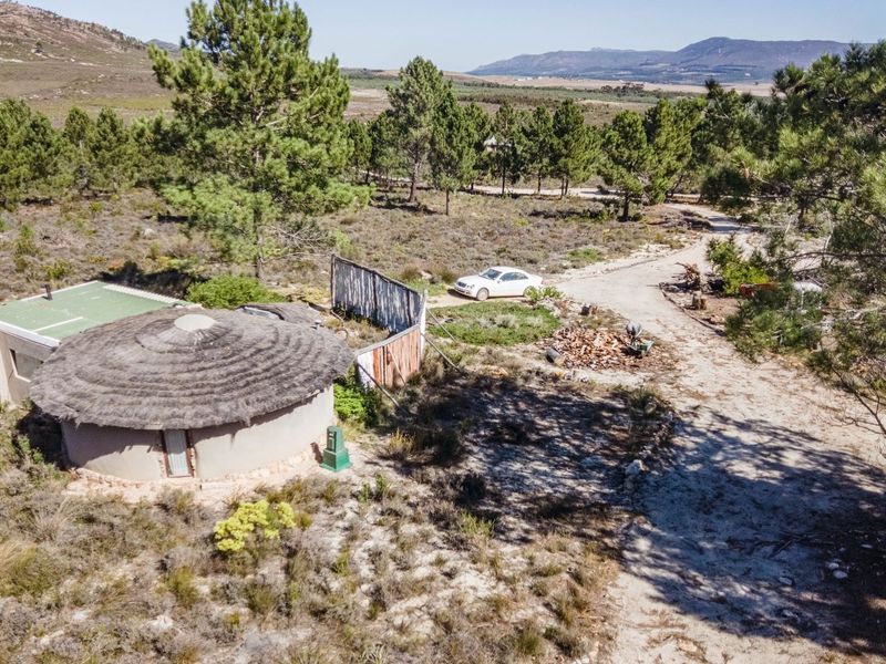 Aerial view of a rural farm property featuring a circular structure with a thatched roof, set within a natural landscape with a gravel driveway.