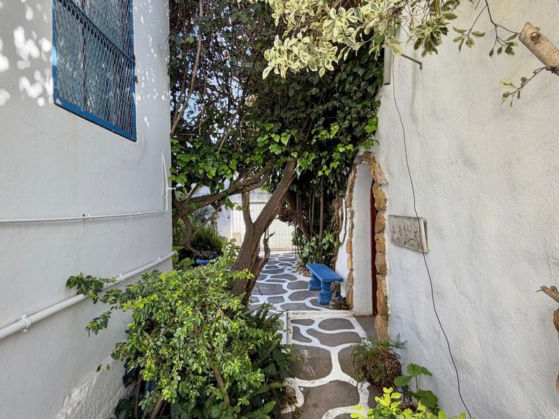 A narrow exterior walkway featuring stone paving, mature greenery, and an arched entrance set against textured white walls.