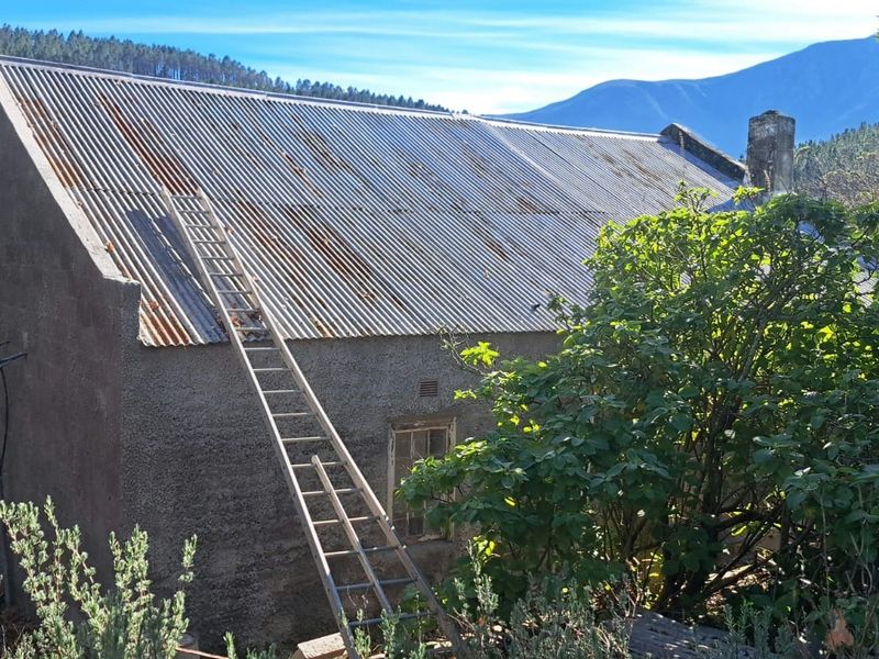 Rear view of a rustic farm building featuring a corrugated metal roof and stone chimney, set against a scenic mountain landscape.