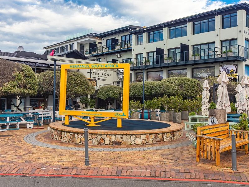 Exterior view of the Hermanus Waterfront commercial complex featuring brick-paved courtyard, stone-clad platform, and multi-story building with balconies.