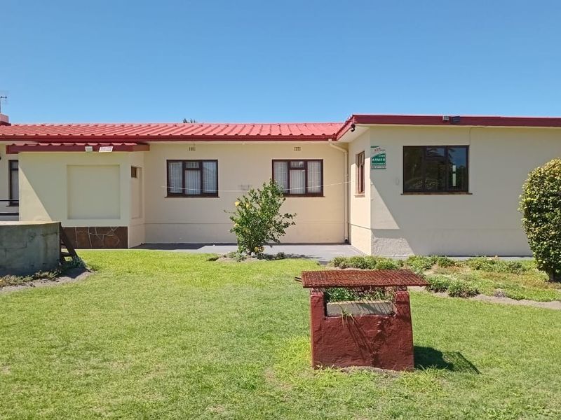 Rear view of the cottage featuring a red corrugated metal roof, cream-painted exterior walls, aluminium windows, a concrete patio, and a built-in braai area.