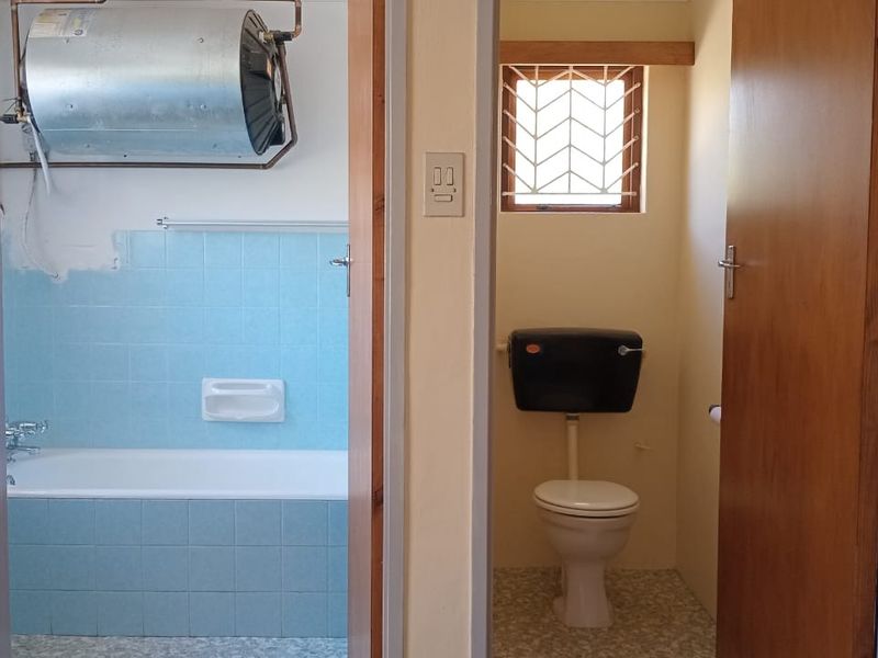 A functional bathroom and separate toilet area featuring a bathtub, tiled splashbacks, and an exposed electric water heater, requiring cosmetic updates.