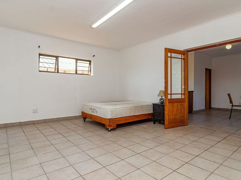 A spacious bedroom featuring tiled flooring, a large window with security bars, and direct access to an adjacent living area through a timber door.