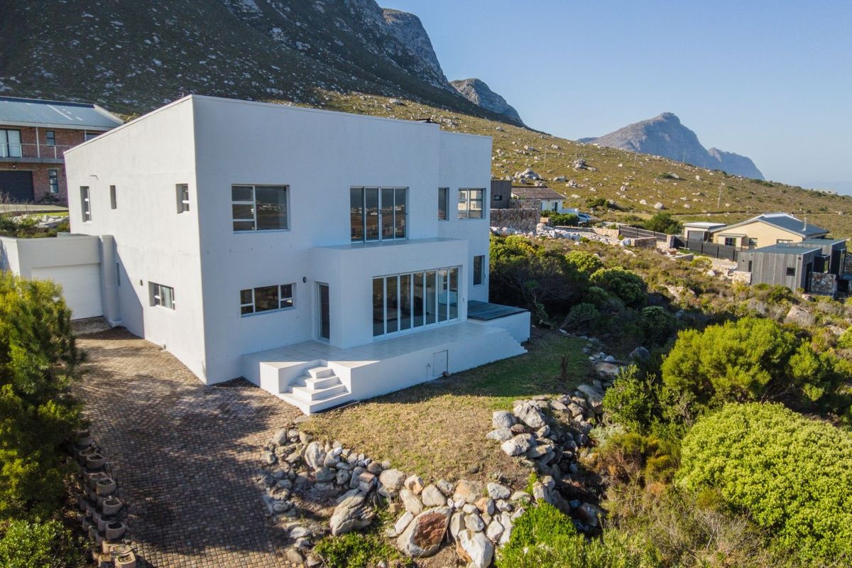 Aerial view of a modern white double-story house featuring a paved driveway, tiled patio, and mountain backdrop in a scenic coastal setting.