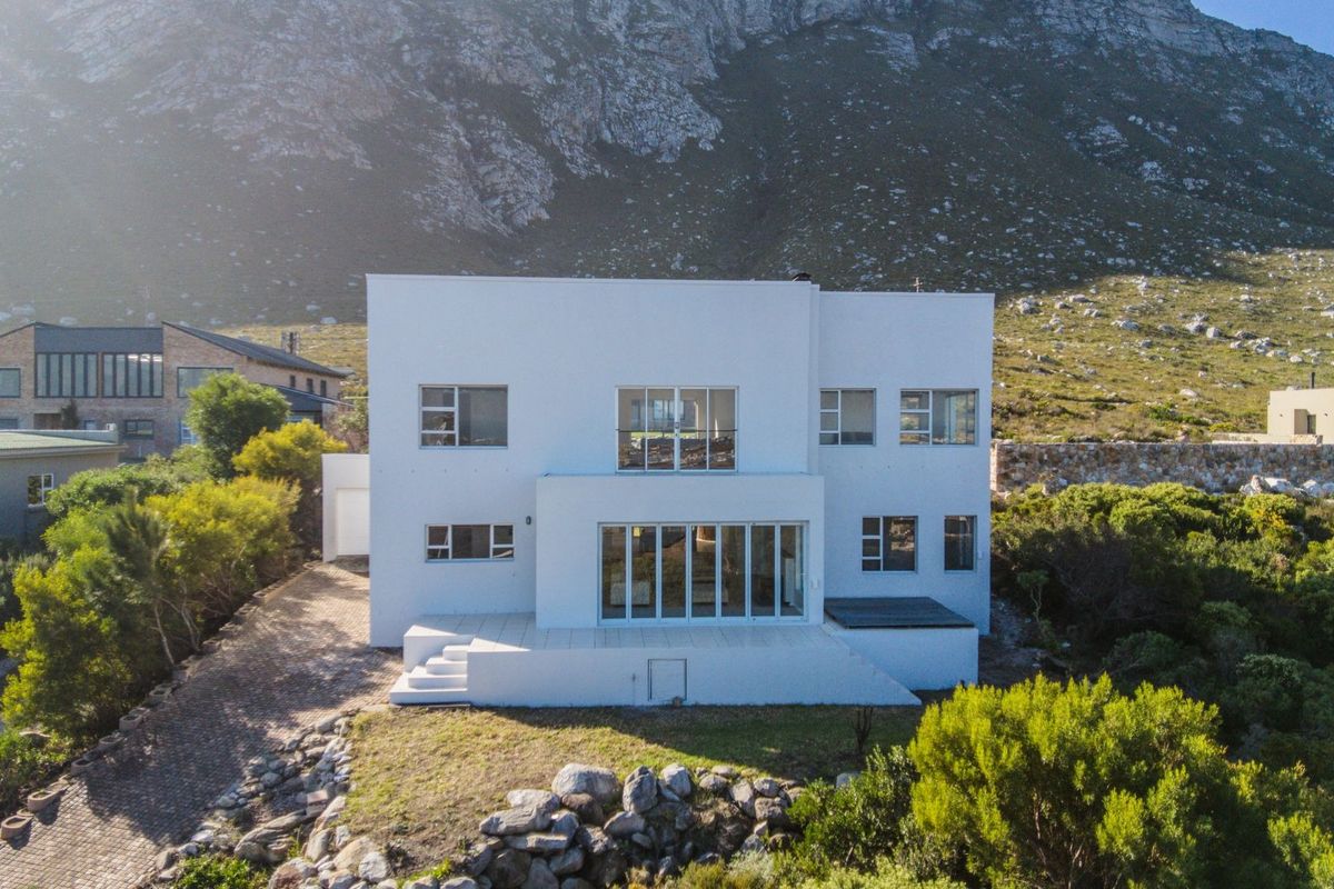 Aerial view of a modern white multi-story residence featuring large glass sliding doors, a raised patio, and a scenic mountain backdrop.