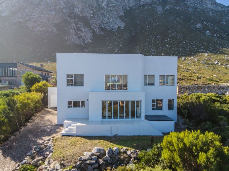 Aerial view of a modern white multi-story residence featuring large glass sliding doors, a raised patio, and a scenic mountain backdrop.