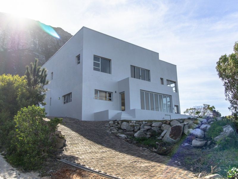 Modern multi-level residential property featuring a clean white facade, paved driveway, and large glass windows set against a natural mountain backdrop.