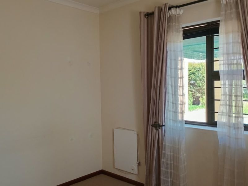 A bright, empty bedroom featuring a wall-mounted heater, neutral walls, and a window with curtains.