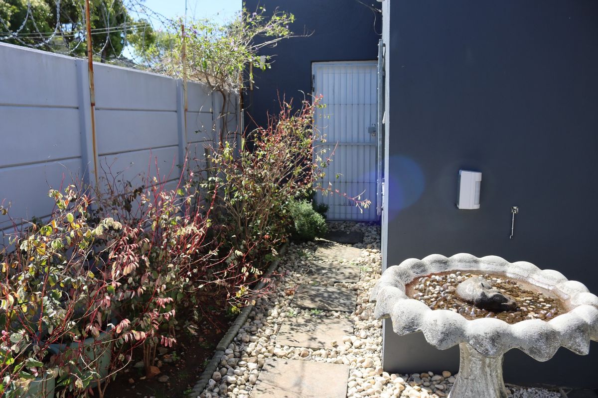 A side garden pathway featuring stone pavers, pebble landscaping, and a secure gate, enclosed by a precast concrete wall with security fencing.