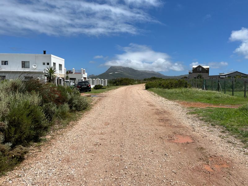A view of an unpaved gravel access road in a residential area, featuring surrounding properties and a scenic mountain backdrop.