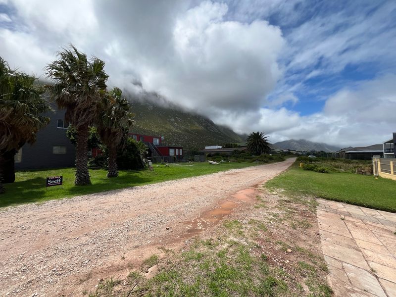 A view of the unpaved street approach to the residential property, featuring surrounding greenery and a scenic mountain backdrop.