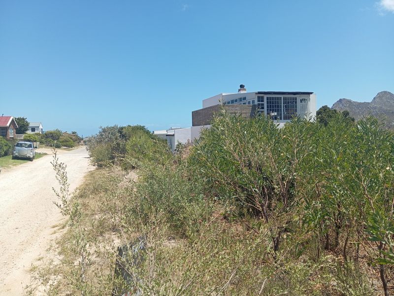 A view of a residential property from an unpaved street, featuring native coastal vegetation and a multi-level dwelling in the background.
