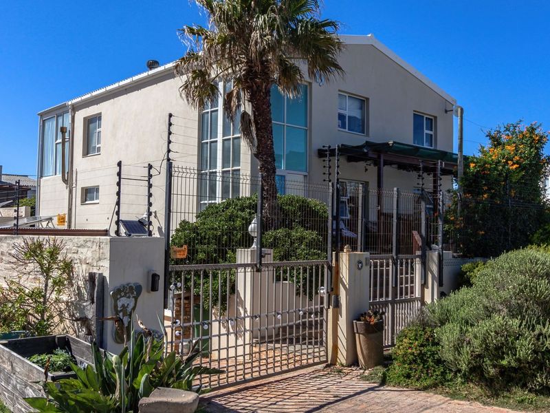 Front exterior view of a two-story house featuring a paved driveway, secure metal gate, electric fencing, and a small garden area.