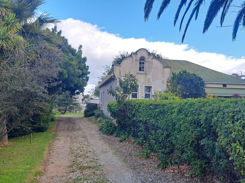 A view of the farm property entrance featuring a gravel driveway, mature landscaping, and the side elevation of the main farmhouse with gabled architecture.