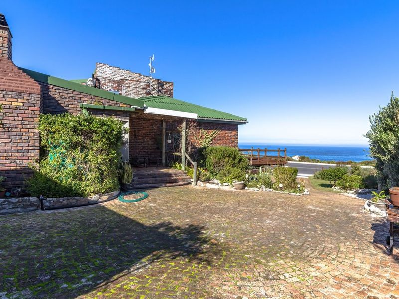 Front exterior view of a brick house featuring a paved driveway, corrugated metal roof, and ocean views in the background.