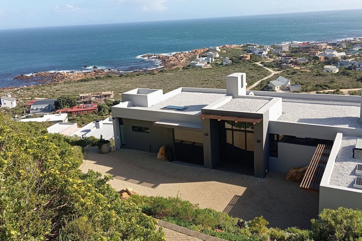 An elevated aerial view of a modern coastal residence featuring a flat roof, solar panels, and a paved driveway with scenic ocean views.