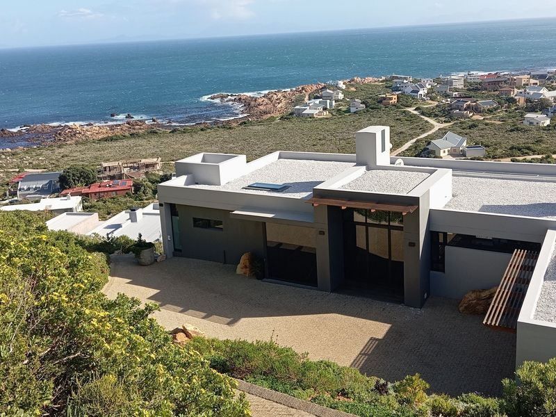 An elevated aerial view of a modern coastal residence featuring a flat roof, solar panels, and a paved driveway with scenic ocean views.