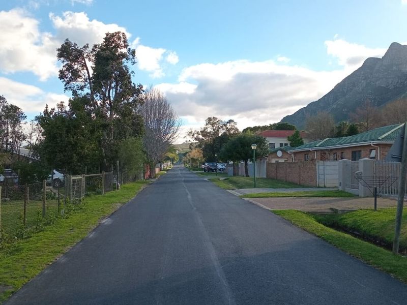 A view of the residential street approach featuring established boundary walls, mature trees, and scenic mountain views in the background.