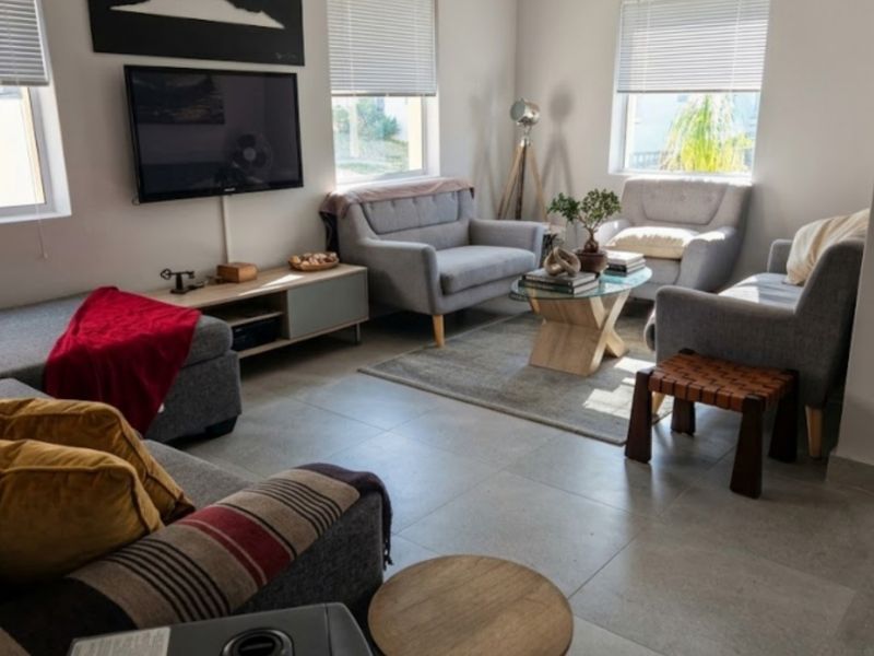 A bright and comfortable living room featuring large windows with venetian blinds and neutral tiled flooring throughout.