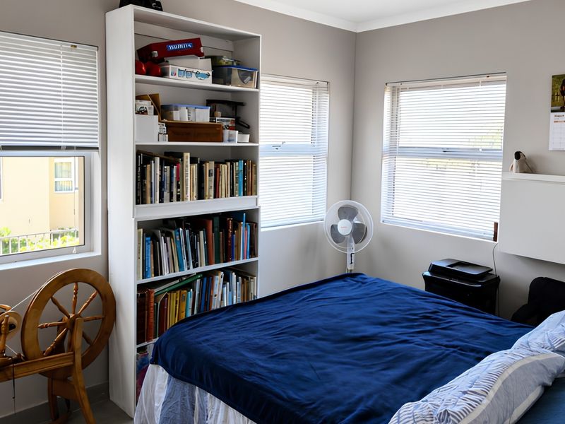 A functional bedroom featuring neutral walls, multiple windows with fitted venetian blinds, and a built-in shelving unit for storage.