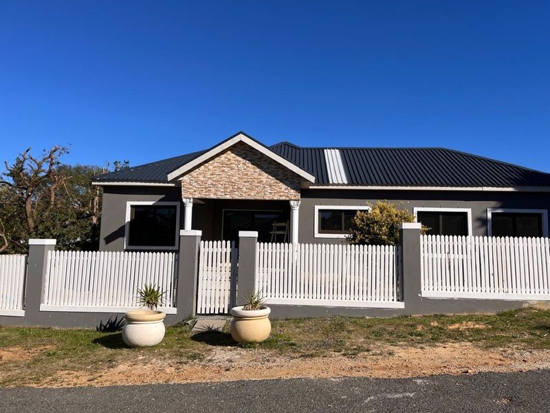 Front elevation of a single-story house featuring a dark metal roof, stone-clad entrance gable, and a white picket fence with concrete pillars.