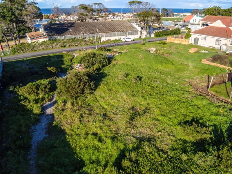 An elevated aerial view of a vacant residential plot featuring natural vegetation, perimeter fencing, and surrounding neighborhood context with distant ocean views.