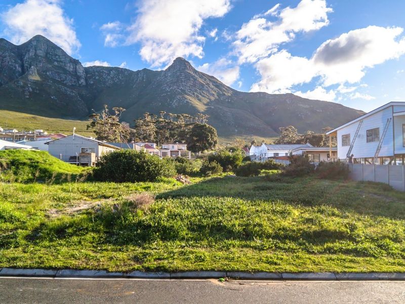 A vacant residential plot of land featuring natural grass cover, situated in a scenic area with mountain views and surrounding residential properties.
