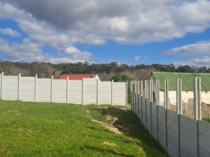 A vacant residential plot featuring a precast concrete boundary wall and grass lawn, with views of an adjacent structure.