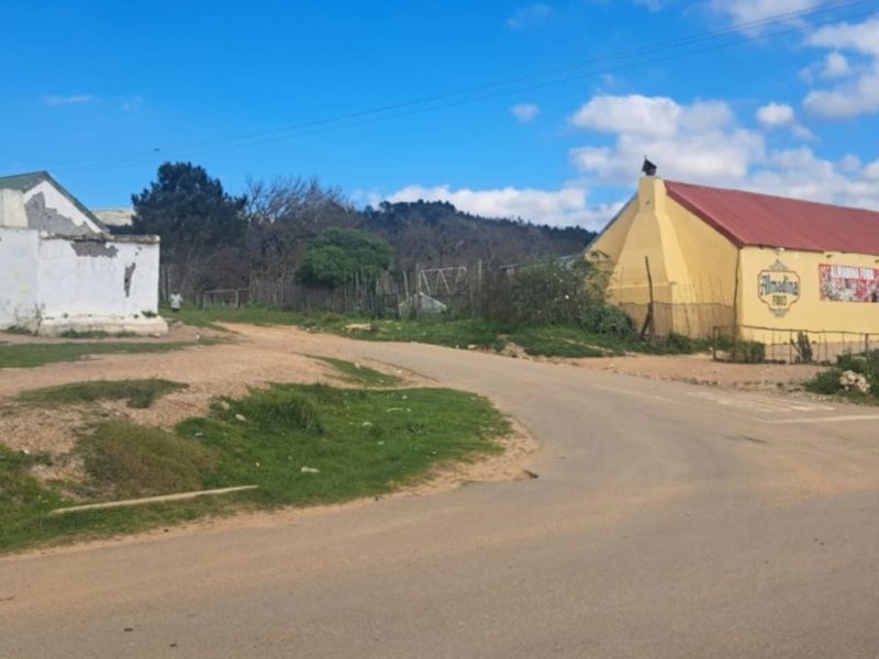 Street view of a residential area featuring an unpaved road, utility infrastructure, and existing structures in varying states of repair.