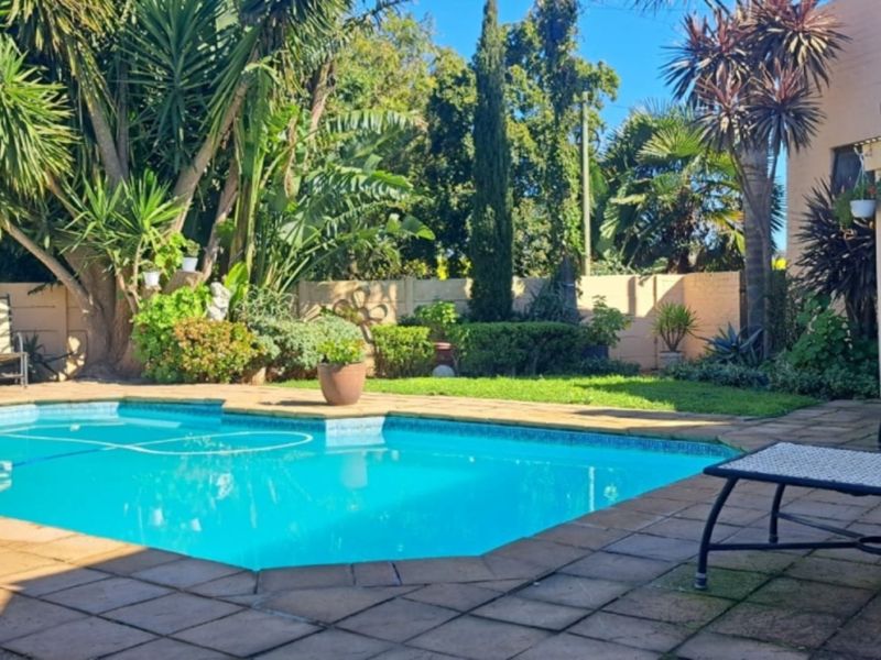 A well-maintained outdoor pool area featuring a paved patio, established garden landscaping, and a covered patio space for outdoor relaxation.