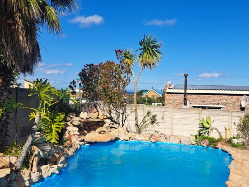 Outdoor pool area featuring a rock water feature, paved surround, and precast concrete boundary wall in a sunny garden setting.