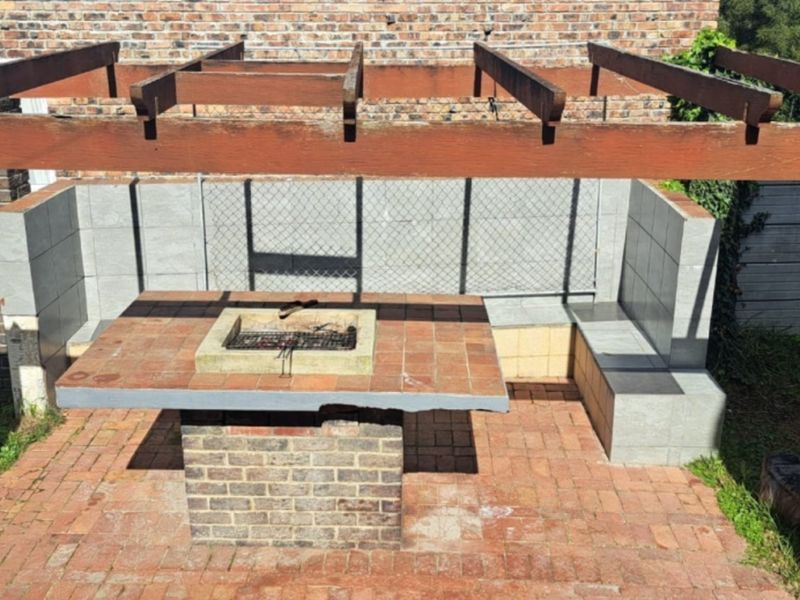 Outdoor entertainment area featuring a brick-built braai, tiled counter, built-in bench seating, and a timber pergola structure set over brick paving.