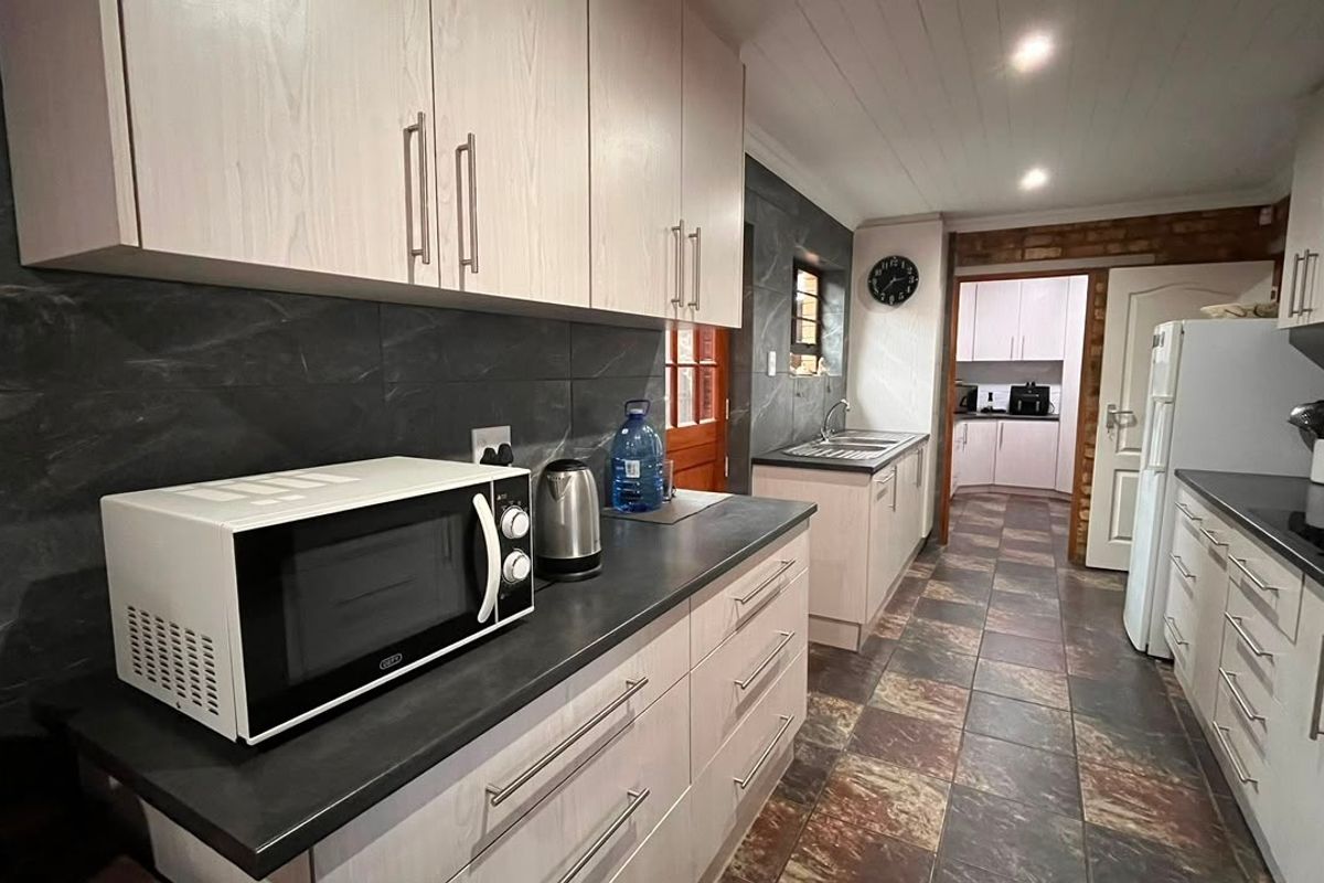 A functional kitchen featuring light-toned cabinetry, dark stone-look countertops, tiled flooring, and a secondary scullery area visible through the doorway.