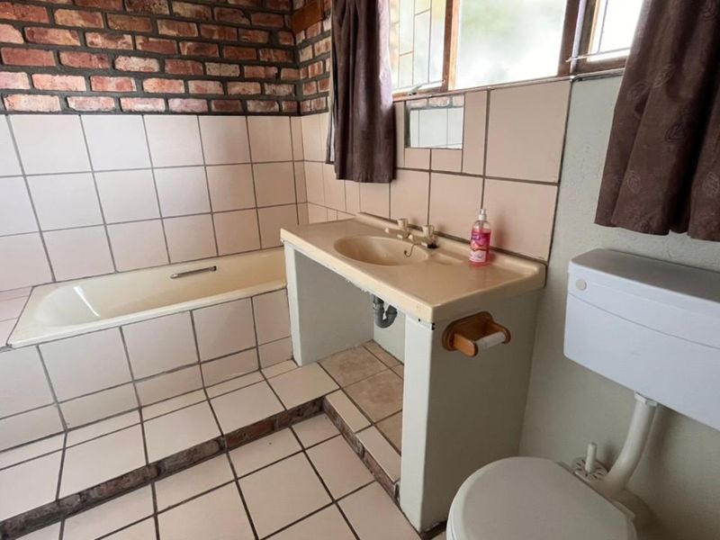 A dated bathroom featuring a built-in bathtub, tiled vanity with integrated basin, and a standard toilet, finished with exposed brick and ceramic wall tiles.