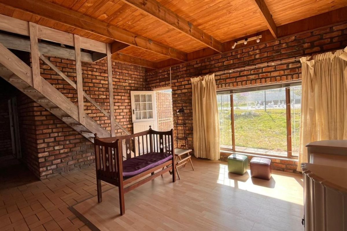 Rustic living area featuring exposed brick walls, timber beam ceiling, and a wooden staircase, with large windows providing natural light and garden views.