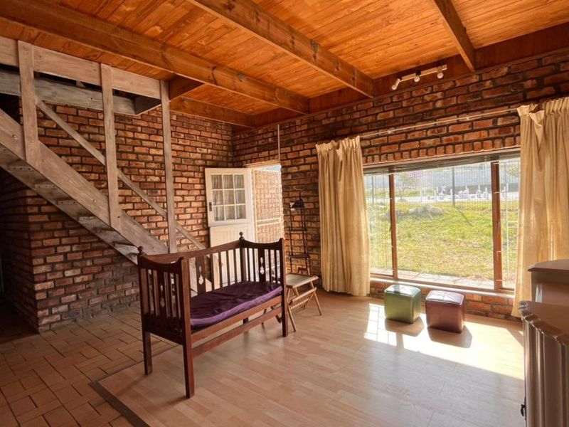 Rustic living area featuring exposed brick walls, timber beam ceiling, and a wooden staircase, with large windows providing natural light and garden views.