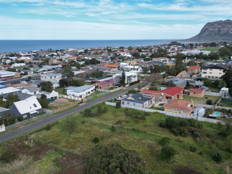 An aerial view of a vacant residential plot situated in a coastal neighborhood with surrounding houses and mountain views.