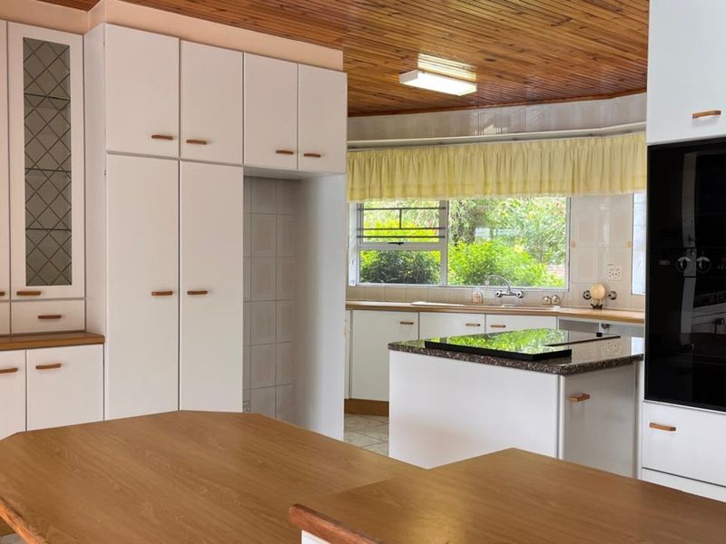 A spacious kitchen featuring white built-in cabinetry, a central island with a hob, built-in oven, and a distinctive timber-paneled ceiling.