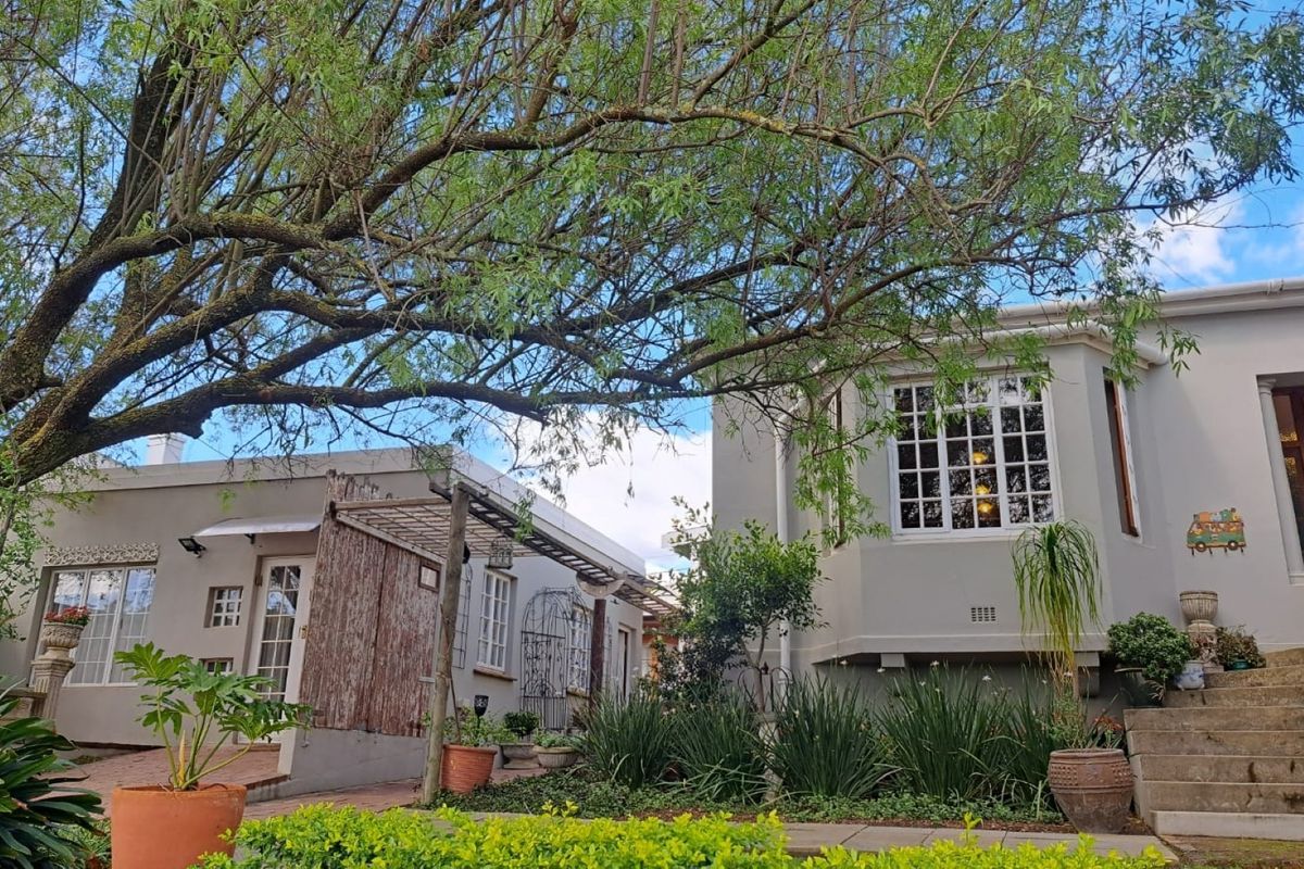 A charming rear exterior view featuring a bay window, paved patio area, pergola, and landscaped garden with concrete steps leading to the entrance.